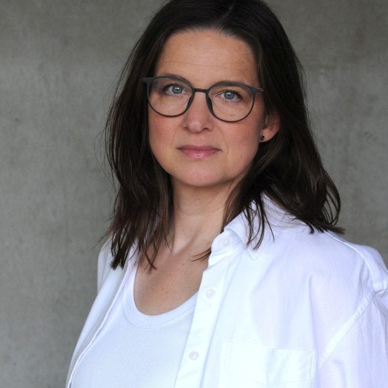 Julia Sittmann, Beraterin und Coach Julia Sittmann lächelt in die Kamera. Im Hintergrund ist ein Wandbild verschwommen zu sehen.
